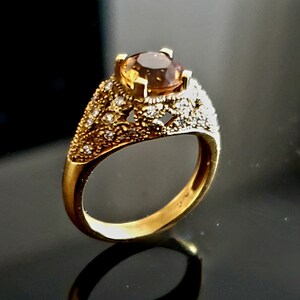 May include: A gold ring with a large, oval-shaped brown gemstone surrounded by smaller, white gemstones. The ring has a delicate, intricate design.