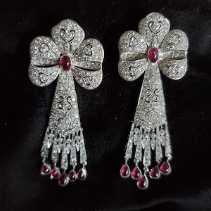 Puede incluir: Un par de pendientes de plata con un diseño en forma de trébol, cada uno con una piedra preciosa roja en el centro y una franja de piedras preciosas más pequeñas.