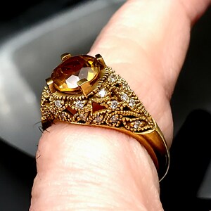 May include: A gold ring with a large, round, yellow gemstone surrounded by smaller, clear gemstones. The ring has a delicate, intricate design.