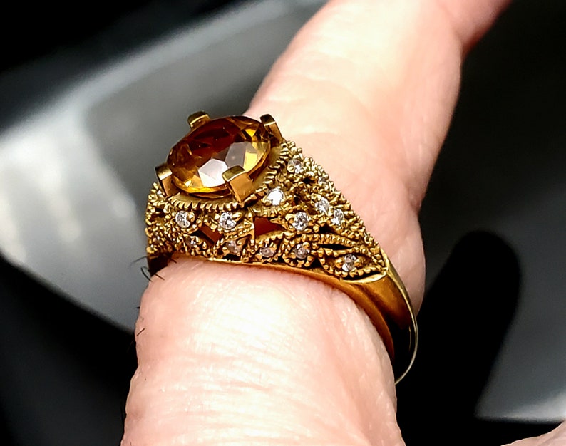 May include: A gold ring with a large, round, yellow gemstone surrounded by smaller, clear gemstones. The ring has a delicate, intricate design.
