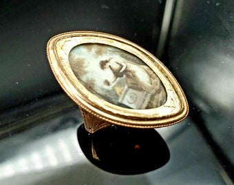 May include: A gold ring with a cameo featuring a woman in a white dress. The cameo is set in a gold oval frame with a beaded edge.