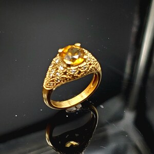 May include: A gold ring with a large, round, yellow gemstone surrounded by smaller, white gemstones. The ring has a detailed, ornate design.