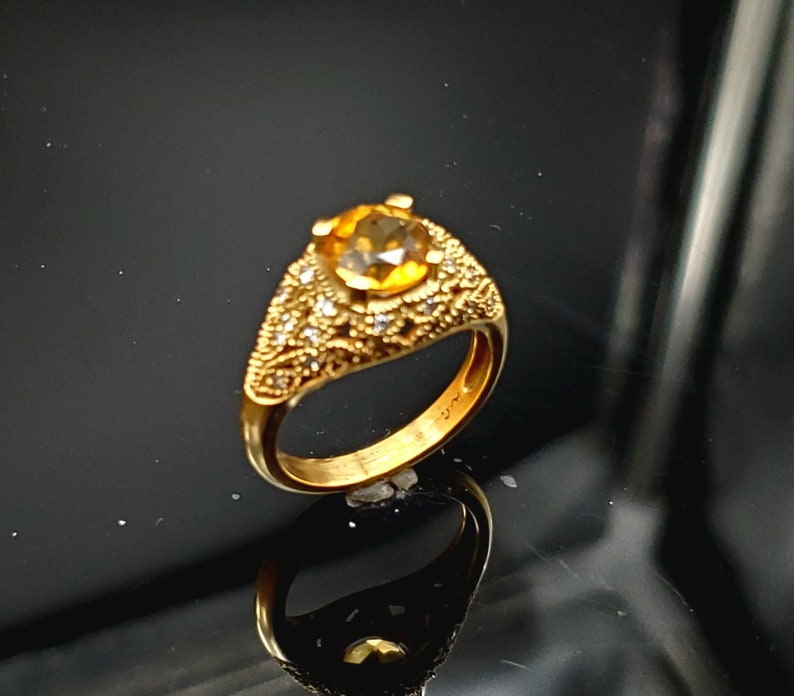 May include: A gold ring with a large, round, yellow gemstone surrounded by smaller, white gemstones. The ring has a detailed, ornate design.