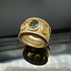 May include: A gold ring featuring three oval-shaped gemstones. The central stone is a dark green, and the two side stones are a light yellow.