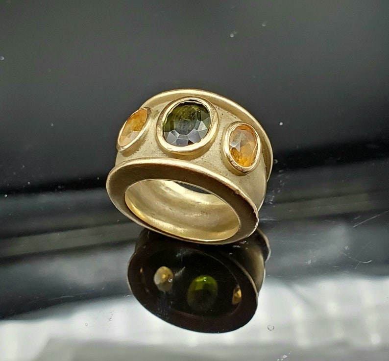 May include: A gold ring featuring three oval-shaped gemstones. The central stone is a dark green, and the two side stones are a light yellow.