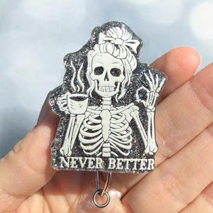 May include: A black glittery badge reel featuring a white skeleton illustration with a messy bun, holding a steaming coffee mug and making an okay hand gesture. The text NEVER BETTER is printed in white at the bottom of the acrylic piece.