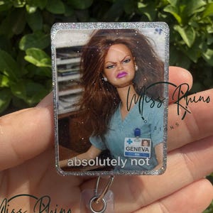 May include: A rectangular badge reel with a glittery border featuring a doll with a displeased expression. The text "absolutely not" is below the image. The badge reel has a silver clip and a glittery button.