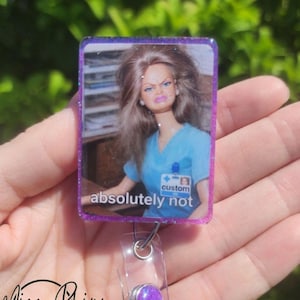 May include: A rectangular badge reel with a purple glitter border features a doll image with the text "absolutely not." The reel has a clear plastic strap and a purple gem button.