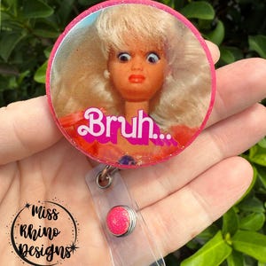 May include: A pink and glittery badge reel with a round design featuring a doll's face with wide eyes and the word "Bruh...". The badge reel has a clear plastic strap and a pink glitter button.