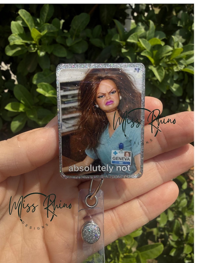 May include: A rectangular badge reel with a glittery border featuring a doll with a displeased expression. The text "absolutely not" is below the image. The badge reel has a silver clip and a glittery button.