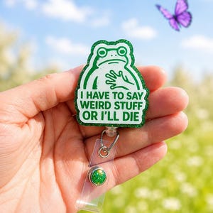 Puede incluir: Un portatarjetas verde con purpurina que presenta una ilustración de una rana blanca con el texto I HAVE TO SAY WEIRD STUFF OR I'LL DIE. El portatarjetas tiene una correa de plástico transparente con un pequeño botón decorativo verde brillante.