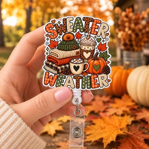 May include: A badge reel featuring a colorful illustration with the text "SWEATER WEATHER". The design includes a stack of folded sweaters, a knit beanie, a coffee cup, a pumpkin, and autumn leaves in shades of orange, green, and brown.