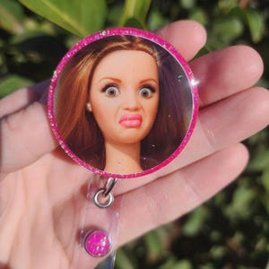 May include: A pink glitter badge reel featuring a doll's face with a surprised expression. The badge reel has a clear plastic strap and a pink glitter button. The doll has red hair, pink lips, and large eyes.
