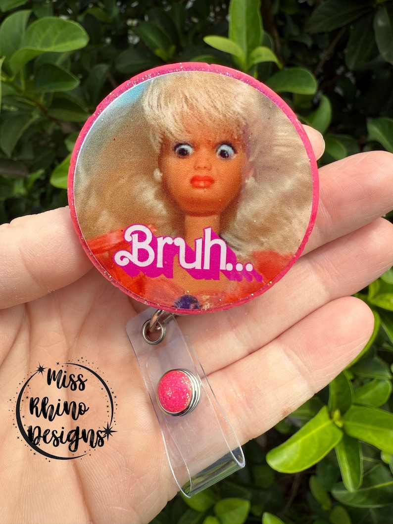 May include: A pink and glittery badge reel with a round design featuring a doll's face with wide eyes and the word "Bruh...". The badge reel has a clear plastic strap and a pink glitter button.