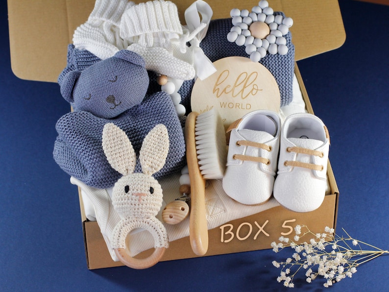 May include: A blue and white baby gift box with a knitted blanket, a stuffed animal, a wooden rattle, a wooden brush, a pair of white baby shoes, and a wooden sign that says "hello world".