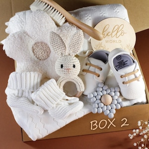 May include: A white gift box filled with baby items, including a knitted blanket, a crocheted bunny rattle, a wooden sign that says "hello world", and a pair of white baby shoes. The box is labeled "Box 2".