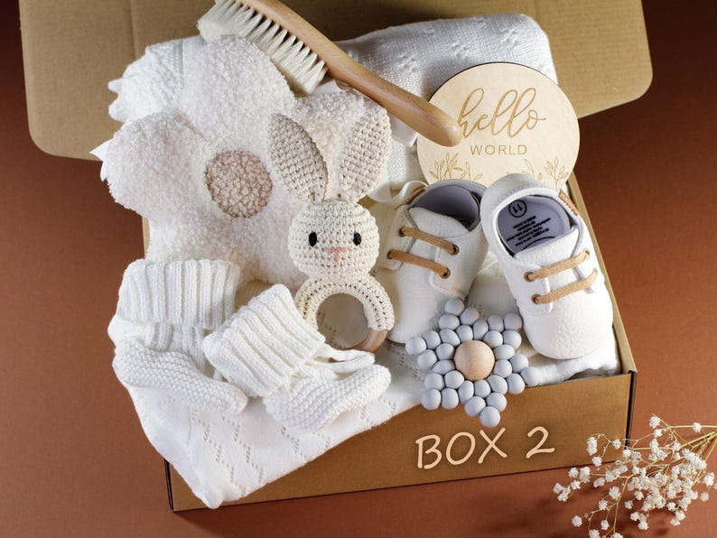 May include: A white gift box filled with baby items, including a knitted blanket, a crocheted bunny rattle, a wooden sign that says "hello world", and a pair of white baby shoes. The box is labeled "Box 2".