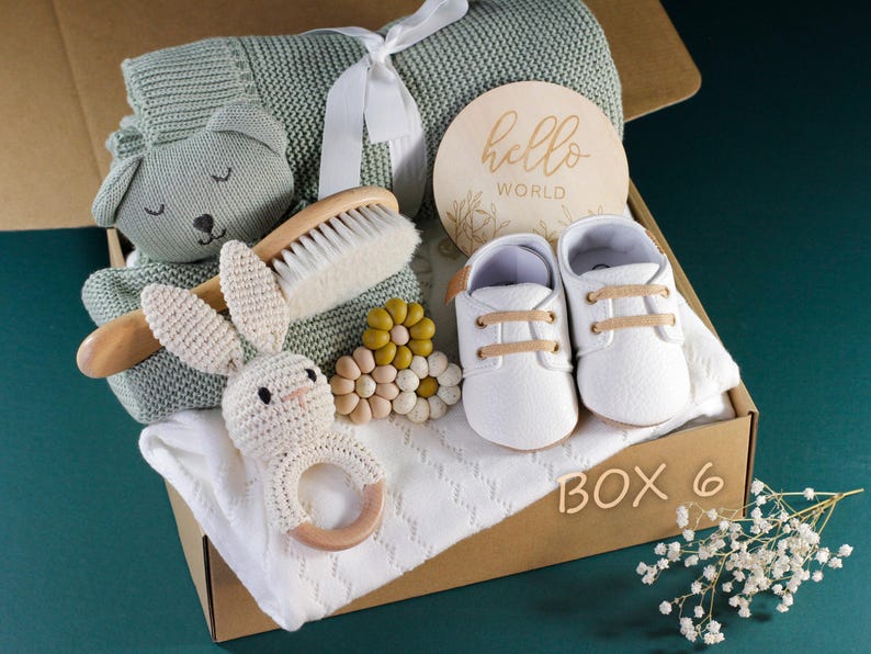 May include: A gift box filled with baby items, including a knitted blanket, a wooden brush, a crocheted bunny rattle, a wooden teether, a pair of white baby shoes, and a wooden sign that says "hello world". The box is labeled "BOX 6".