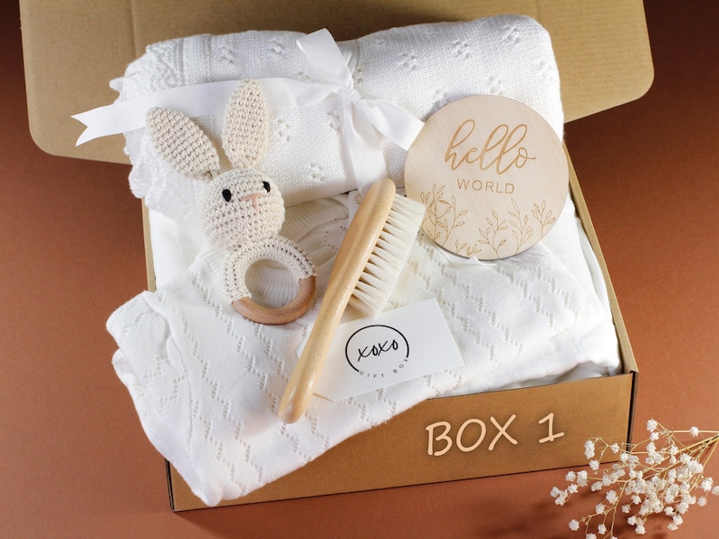 May include: A white gift box filled with baby items, including a knitted blanket, a wooden rattle, a wooden hair brush, and a wooden sign that says "hello world".