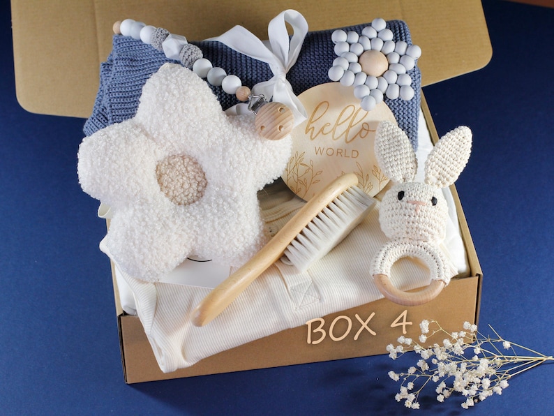 May include: A gift box filled with baby items, including a knitted blanket, a flower-shaped plush toy, a wooden rattle, a wooden hairbrush, a crocheted bunny toy, and a wooden sign that says "hello world".
