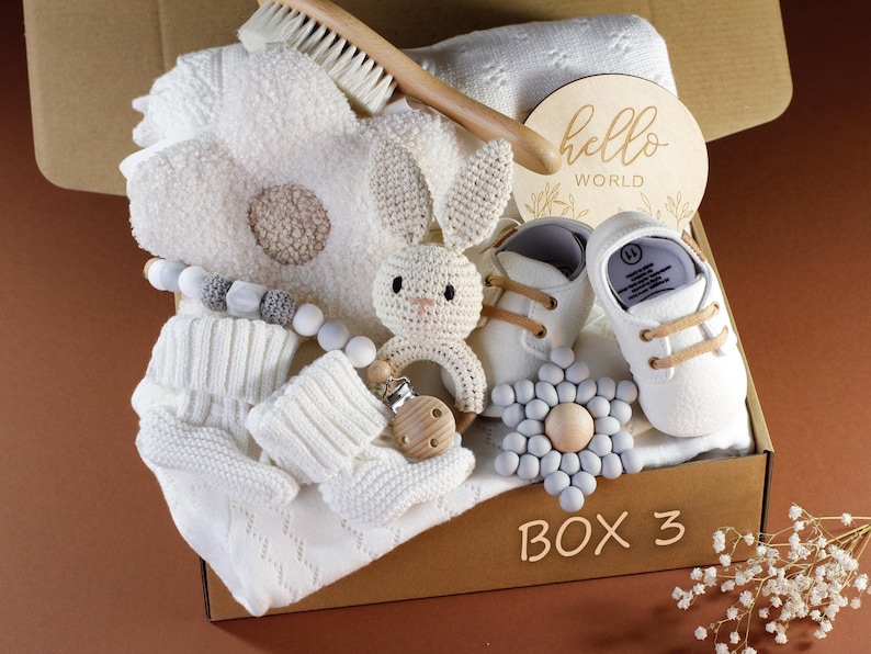 May include: A brown cardboard box filled with baby items, including a white knitted blanket, a white crocheted bunny rattle, a wooden teether, a pair of white baby shoes, a white knitted hat, a wooden sign that says "hello world", and a gray and white flower-shaped teether. The box is labeled "BOX 3".