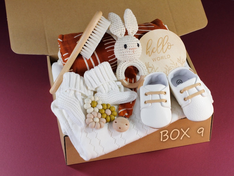 May include: A brown cardboard box with the words "BOX 9" printed on the front. The box is filled with baby items, including a white knitted sweater, a pair of white baby shoes, a wooden bunny rattle, a wooden brush, and a wooden sign that says "hello world".