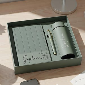 Könnte beinhalten: Ein salbeigrünes Geschenkset mit Notizbuch, Stift und Isolierflasche, alle mit dem Namen "Sophia" beschriftet. Das Set wird in einer passenden Box präsentiert. Eine kleine Karte mit dem Namen "Sophia" und floralen Akzenten ist ebenfalls enthalten.