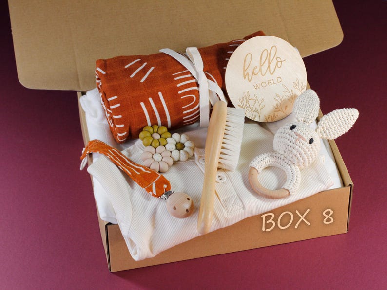 May include: A brown cardboard box filled with baby items, including a white onesie, a wooden brush, a crocheted bunny rattle, a wooden teething ring, a muslin swaddle blanket with a geometric pattern, a wooden teether, and a wooden sign that says "hello world". The box is labeled "BOX 8".