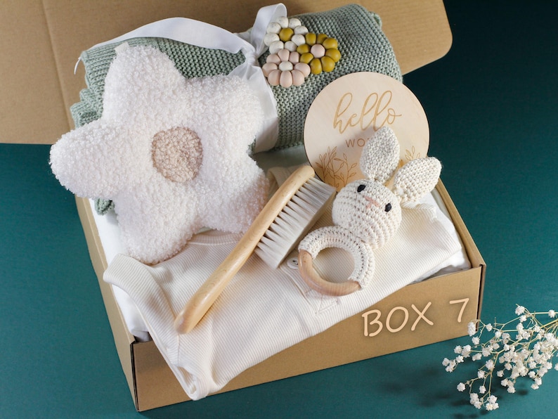 May include: A brown cardboard box filled with baby items, including a white knitted bunny rattle, a wooden brush, a white knitted flower, a white onesie, a green knitted blanket with a flower applique, and a wooden sign that says "hello world". The box is labeled "Box 7".