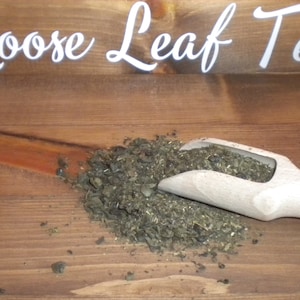 Könnte beinhalten: Ein Holzlöffel gefüllt mit losen Blatttee liegt auf einer Holzoberfläche. Der Tee ist dunkelgrün und scheint eine Mischung aus Kräutern und Gewürzen zu sein. Die Worte "Loose Leaf Tea" sind in Weiß auf einem Holzschild über dem Tee geschrieben.