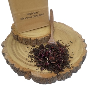 May include: A brown paper bag labelled 'Wild Cherry' Black Tea & Cherry Blend, accompanied by a wooden spoon filled with a loose-leaf tea mixture containing dark tea leaves, dried red cherry pieces, and purple floral elements.