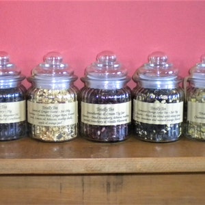 May include: Ten glass jars filled with loose leaf tea, each with a label that reads "Buddy Tea" and a description of the tea blend. The jars are arranged in a row on a wooden shelf.