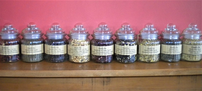 May include: Ten glass jars filled with loose leaf tea, each with a label that reads "Buddy Tea" and a description of the tea blend. The jars are arranged in a row on a wooden shelf.