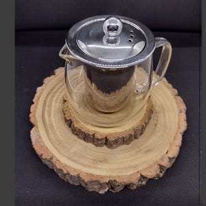 Puede incluir: Una tetera de vidrio transparente con tapa e infusor de acero inoxidable se encuentra sobre una base de madera. La tetera tiene un asa y un pico.