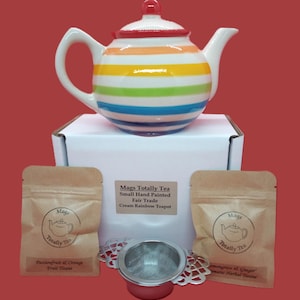 Könnte beinhalten: Eine handbemalte cremefarbene Teekanne mit Regenbogenstreifen, einem roten Deckel und einem weißen Griff und Ausguss. Das Set enthält zwei Teebeutel, einen Tee-Infuser und eine Box. Der Text auf der Box lautet "Mags Totally Tea Cream Rainbow Teapot."