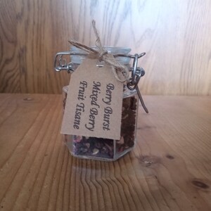 May include: A glass jar with a metal clasp and a brown paper tag that reads "Berry Burst Mixed Berry Fruit Tisane". The jar is filled with dried fruit and herbs.