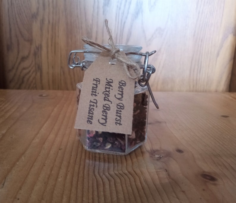 May include: A glass jar with a metal clasp and a brown paper tag that reads "Berry Burst Mixed Berry Fruit Tisane". The jar is filled with dried fruit and herbs.