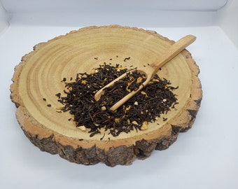 May include: A wooden bowl filled with loose leaf black tea with orange peel pieces. A wooden spoon is resting on top of the tea.