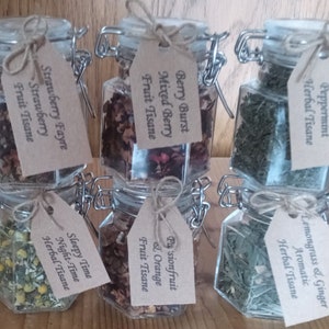 May include: Six glass jars with metal lids and twine tied around the lids. Each jar contains dried herbs or fruit and has a brown paper tag with a handwritten label. The labels read: "Totally Mint Peppermint Herbal Tisane", "Lemongrass & Ginger Aromatic Herbal Tisane", "Berry Burst Mixed Berry Fruit Tisane", "Pasionfruit & Orange Fruit Tisane", "Sleepy Time Night-Time Herbal Tisane", and "Strawberry Fayre Strawberry Fruit Tisane".