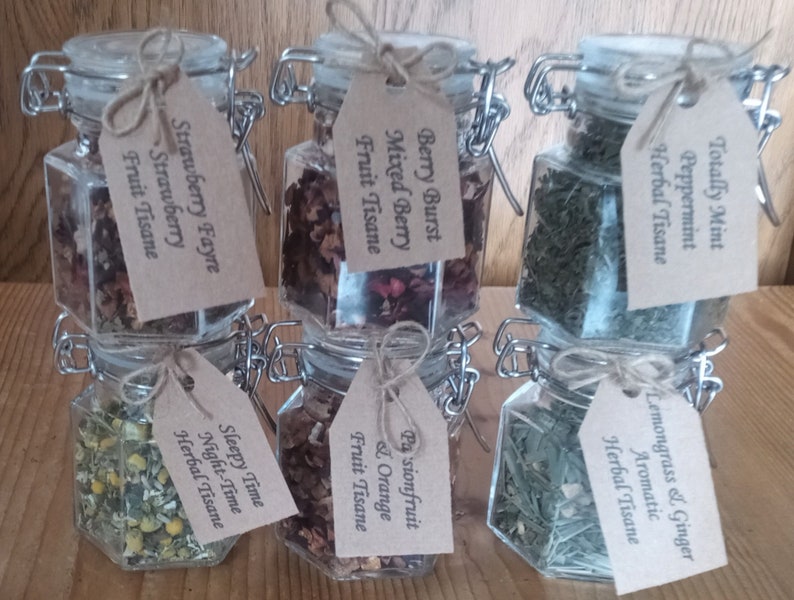 May include: Six glass jars with metal lids and twine tied around the lids. Each jar contains dried herbs or fruit and has a brown paper tag with a handwritten label. The labels read: "Totally Mint Peppermint Herbal Tisane", "Lemongrass & Ginger Aromatic Herbal Tisane", "Berry Burst Mixed Berry Fruit Tisane", "Pasionfruit & Orange Fruit Tisane", "Sleepy Time Night-Time Herbal Tisane", and "Strawberry Fayre Strawberry Fruit Tisane".