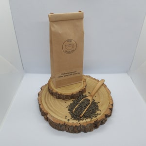 May include: A brown paper bag with the text "Mags Really Tea" and "Pinhead Gunpowder Chinese Gunpowder Green Tea" printed on it. The bag is sitting on a wooden platform with a wooden spoon and loose green tea leaves.