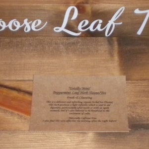 May include: A brown paper card with the text "Totally Mint Peppermint Leaf Herb Tea" and "Fresh & Cleansing" printed on it. The card is on a wooden surface with the text "Loose Leaf Tea" printed on the surface.