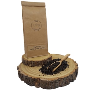 May include: A brown paper bag of High Society Earl Grey loose leaf tea with a Mags Totally Tea logo, placed on a wooden slice next to a small wooden scoop filled with dark tea leaves. The bag features a circular logo with a teapot illustration.