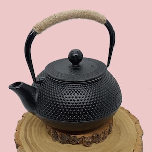 Puede incluir: Tetera de hierro fundido negro con una superficie texturizada y un asa envuelta en cuerda natural. La tetera se exhibe sobre una rodaja de madera, destacando su diseño tradicional. La tapa tiene un pequeño pomo.