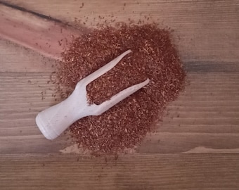 May include: A wooden scoop rests in a pile of reddish-brown loose tea leaves on a wooden surface. The scoop is light-colored and has a forked end. The tea leaves are scattered around the scoop, creating a textured appearance.