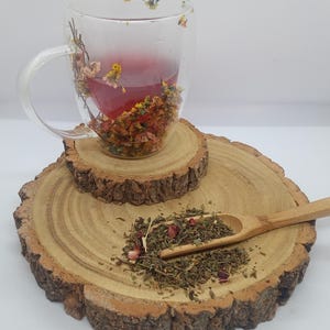 Peut inclure: Une tasse en verre transparente remplie d'un liquide rouge et de fleurs séchées, posée sur un dessous de verre en bois. Une cuillère en bois contient des feuilles de thé en vrac. La tasse a une anse et est ornée de motifs floraux.
