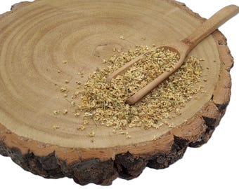 May include: A small wooden scoop rests on a round, natural wood slice, containing a pile of dried, light beige elderflower blossoms. The wood slice features visible growth rings and a rough, dark brown bark edge, providing a textured base.