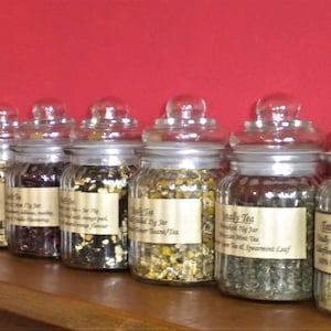 May include: A row of ten glass jars filled with loose leaf tea. The jars have white lids and labels with the text "Totally Tea" and the type of tea inside.