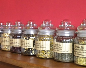 May include: A row of ten glass jars filled with loose leaf tea. The jars have white lids and labels with the text "Totally Tea" and the type of tea inside.
