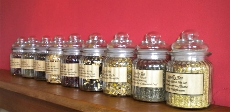 May include: A row of ten glass jars filled with loose leaf tea. The jars have white lids and labels with the text "Totally Tea" and the type of tea inside.